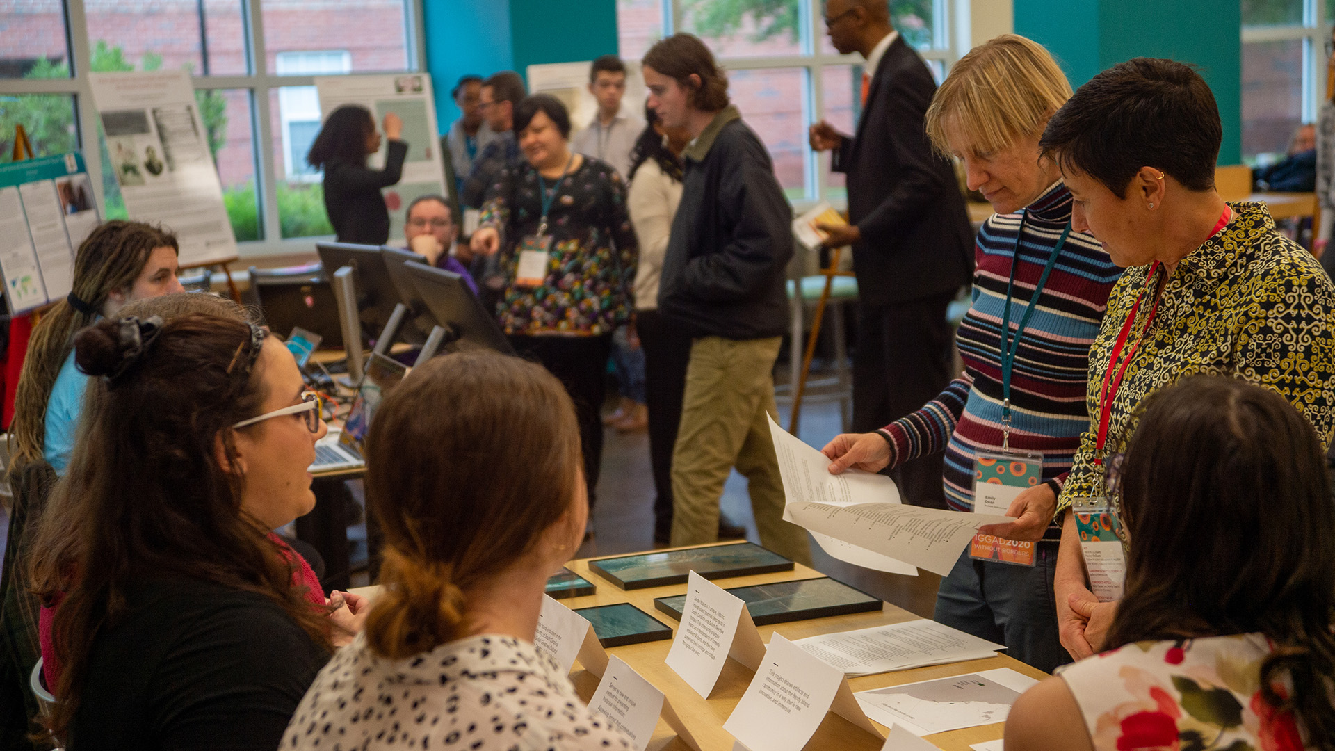 Participants stand around tables at an academic poster session, reviewing printed research materials and discussing projects displayed on boards and tablets.